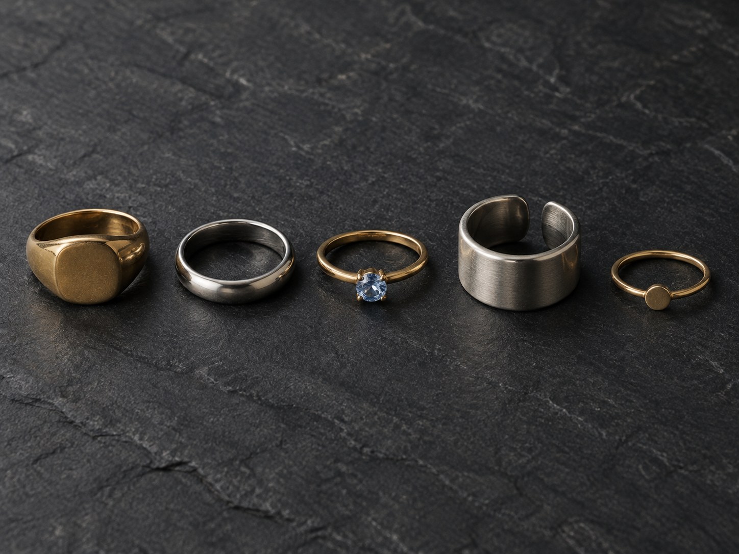 Five distinct rings in a row on dark slate: signet, band, stone ring, thumb ring and pinky ring, AI-generated illustration