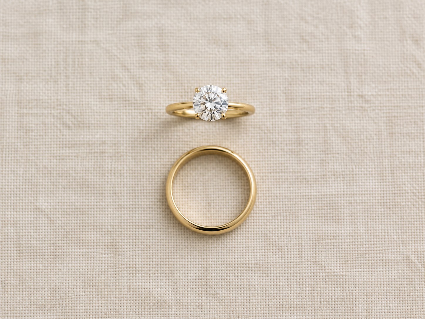 A gold wedding band placed below a solitaire engagement ring showing stacking order on linen, AI-generated illustration