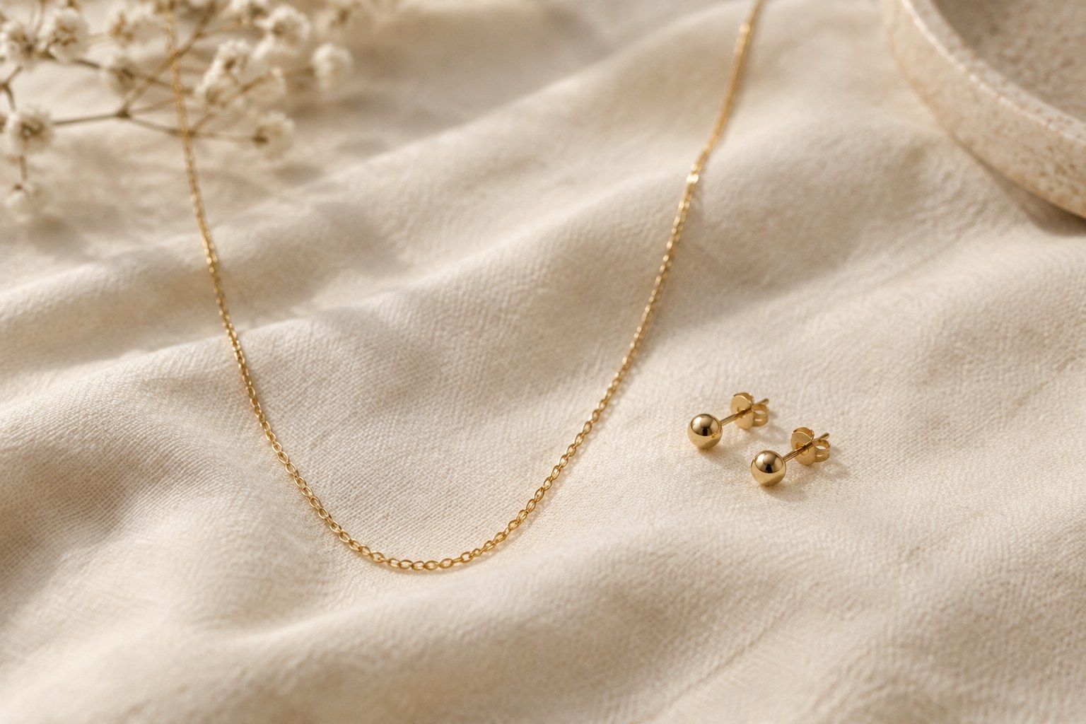 Illustration: small gold stud earrings and a fine chain as an everyday jewellery uniform on cream fabric